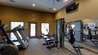 This fitness center is a single-floor facility designed to support a comprehensive workout experience. The space is finished with warm golden-tan walls and durable carpeted flooring, creating a welcoming yet functional atmosphere. Recessed lighting illuminates the room evenly, complemented by natural light streaming in through multiple glass-paned doors and windows along the front wall, giving the space an open and airy feel. A full-length mirror runs along one wall, visually expanding the room and allowing users to monitor their form during workouts. The facility is outfitted with a solid selection of exercise equipment, including cardio machines such as a treadmill and a stationary recumbent bike, as well as a multi-station cable strength training machine and a weight bench with free weights. Wall-mounted flat-screen televisions are positioned for easy viewing from multiple workout stations, enhancing the exercise experience. A decorative clock and informational signage near the entrance add functional touches to the space. The ceiling features a fan for additional air circulation, ensuring comfort during intense workouts. Overall, this fitness center offers a practical and well-appointed environment suited for both cardio and strength training routines.