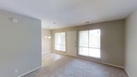 This single-floor space offers a clean, open layout designed for comfortable everyday living. The main living and dining area is bathed in natural light, thanks to multiple large windows fitted with blinds that allow for adjustable brightness throughout the day. A front entry door is centrally positioned, flanked by windows that enhance the sense of openness and connectivity to the outdoors. The room features soft, wall-to-wall carpeting in a neutral tone that adds warmth underfoot and complements the light beige walls throughout. A decorative chandelier hangs gracefully above the dining area, providing an elegant focal point and ambient lighting. The smooth white ceiling with recessed vents contributes to the clean, uncluttered aesthetic of the space. The open-plan design between the living and dining zones creates a seamless flow, making the area feel spacious and versatile. The neutral color palette of beige walls, light carpet, and white trim gives the space a timeless, welcoming character that is easy to personalize. Overall, the space presents a bright, airy, and well-lit environment ideal for both relaxation and entertaining.