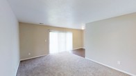 This single-floor residential space offers a clean, functional layout designed for comfortable everyday living. The main living area is the focal point of the unit, characterized by warm beige walls and a textured ceiling that adds subtle depth to the room. Soft, wall-to-wall carpeting covers the living space, providing warmth underfoot, while a transition to hardwood-style flooring is visible toward the perimeter, adding a touch of contrast and visual interest. A large sliding glass door dressed with vertical blinds serves as the primary source of natural light, flooding the room with brightness and offering direct access to the outdoors. The door's generous size creates an open, airy ambiance and provides an easy connection between the interior and exterior. Recessed ventilation is integrated into the ceiling, ensuring efficient climate control throughout the space. White baseboards and trim provide a crisp finish against the neutral walls, contributing to the overall clean and well-maintained aesthetic. The open, uncluttered layout of the living area allows for flexible furniture arrangement and makes the space feel welcoming and adaptable to a variety of interior styles.
