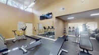 Located at 4200 Community Drive in West Palm Beach, Florida, this fitness center at Azalea Village Apartments is a well-equipped, single-level facility designed to support a well-rounded workout routine. The space is characterized by soaring vaulted ceilings with exposed white structural beams, warm beige walls, and durable rubber flooring throughout, creating a clean and functional environment for exercise.

The main workout area is generously proportioned and filled with a variety of cardio equipment, including treadmills, elliptical trainers, and stationary bikes from True Fitness and Cybex. A dedicated strength training section features adjustable weight benches, dumbbells, medicine balls on a rack, stability balls, and additional cable machines. Large wall-mounted mirrors expand the visual depth of the space and allow users to monitor their form during exercise.

Dual flat-screen televisions are mounted prominently on the wall, providing entertainment during workouts. Natural light filters in through high clerestory windows, complementing the overhead fluorescent lighting. A water fountain is conveniently located near the entrance, and posted fitness center rules ensure a well-maintained environment. Glass doors offer views of the surrounding greenery, connecting the interior to the lush Florida landscape outside.
