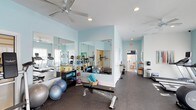 Located at 505 Vorils Ln in Wilmington, NC, this fitness center offers a comprehensive and well-appointed single-floor workout facility designed to support a wide range of exercise routines. The space is bathed in a light, airy ambiance, with soft aqua-toned walls and recessed lighting complemented by a ceiling fan that helps maintain a comfortable environment during workouts. Large wall-to-wall mirrors line key areas of the gym, creating a sense of expansiveness while allowing members to monitor their form during exercise. The flooring throughout is a durable rubber surface, providing both safety and support for high-impact activities. The facility is thoughtfully organized into distinct workout zones. One area is dedicated to strength and flexibility training, equipped with a weight bench, stability balls, resistance tools, medicine ball racks, and foam rollers. A separate section houses cardio equipment, including treadmills and other aerobic machines, visible along the perimeter. A hallway connects to additional amenity spaces, suggesting access to restrooms or other support areas. Natural light filters in through windows, enhancing the bright and motivating atmosphere. The overall design is clean, functional, and welcoming, making this fitness center a well-rounded destination for residents and users seeking a convenient and fully equipped place to exercise.