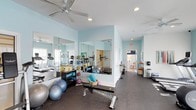 Located at 505 Vorils Ln in Wilmington, NC, this fitness center offers a comprehensive and well-appointed single-floor workout facility designed to support a wide range of exercise routines. The space is bathed in a light, airy ambiance, with soft aqua-toned walls and recessed lighting complemented by a ceiling fan that helps maintain a comfortable environment during workouts. Large wall-to-wall mirrors line key areas of the gym, creating a sense of expansiveness while allowing members to monitor their form during exercise. The flooring throughout is a durable rubber surface, providing both safety and support for high-impact activities. The facility is thoughtfully organized into distinct workout zones. One area is dedicated to strength and flexibility training, equipped with a weight bench, stability balls, resistance tools, medicine ball racks, and foam rollers. A separate section houses cardio equipment, including treadmills and other aerobic machines, visible along the perimeter. A hallway connects to additional amenity spaces, suggesting access to restrooms or other support areas. Natural light filters in through windows, enhancing the bright and motivating atmosphere. The overall design is clean, functional, and welcoming, making this fitness center a well-rounded destination for residents and users seeking a convenient and fully equipped place to exercise.
