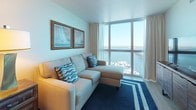 This waterfront residence is thoughtfully designed to maximize its stunning surroundings, with floor-to-ceiling windows that frame panoramic views of the marina and river throughout the main living spaces. The open layout connects the living area and bedroom seamlessly, both oriented to take full advantage of the sweeping waterfront vistas.

The living area is styled with a coastal aesthetic, featuring a plush sectional sofa in neutral tones accented with nautical-themed throw pillows and framed sailboat artwork. A coordinating accent chair and warm lamp complete the inviting seating arrangement, all anchored by a bold navy striped area rug.

The bedroom continues the waterfront theme, offering direct water views through expansive corner windows. Rich dark-wood furniture, including a bed frame, dresser, and nightstand, contrasts elegantly with the light, neutral carpet and soft blue bedding. Framed botanical and coastal artwork adorns the walls, reinforcing the serene ambiance.

The kitchen is efficiently appointed with warm wood cabinetry, granite-style countertops, stainless steel appliances including a full-size refrigerator and built-in microwave, a cooktop, and a sink — providing all the essentials for comfortable in-unit living.

The bathroom features light tile flooring, wood-toned vanity cabinetry, and a clean, functional layout with ample storage.