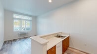 Located at 12495 SW 172nd Terrace in Beaverton, Oregon, this two-bedroom, one-bathroom residence offers a practical and well-appointed single-floor layout. The open-concept living and kitchen area forms the heart of the home, where light wood-style flooring, recessed lighting, and crisp white walls create a bright, clean aesthetic. The kitchen features warm-toned wood cabinetry, light countertops with a peninsula that extends into the living area, a stainless steel sink, white appliances including a refrigerator and range, and ample upper cabinet storage. A wall-mounted HVAC unit and a large window with vertical blinds in the living area allow for natural light and climate control. Both bedrooms are carpeted in a neutral grey tone and feature white walls, natural light from windows with vertical blinds, and notable ceiling height. The primary bedroom includes a built-in closet with shelving, while the secondary bedroom is compact and efficiently designed. A hallway connects the rooms to the full bathroom, which is equipped with wood-tone cabinetry, a wide vanity mirror, wood-style flooring, and a tub-shower combination fitted with accessibility grab bars. The overall design is modern, clean, and functional.