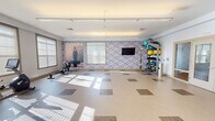 Located at 8085 Sandpiper Cir in Nottingham, MD, this well-appointed fitness facility offers a bright, open layout designed to support a wide range of workout styles. The single-floor space is finished with durable, light-toned flooring accented by subtle patterned tile inserts, providing a clean and modern aesthetic throughout.

The main exercise area is generously proportioned, allowing ample room for both equipment use and open-floor activities such as stretching or group fitness. Natural light streams in through multiple windows fitted with slatted blinds, creating a well-lit environment that enhances the sense of openness. Overhead linear lighting fixtures further illuminate the space evenly.

Cardio equipment, including a treadmill and recumbent bike, is positioned near the windows, while a rowing machine occupies the open-floor zone. A full-length mirrored wall with a ballet barre defines a dedicated studio section, ideal for yoga, dance, or balance training. This area is reflected back through the mirrors, visually expanding the space.

A prominent multi-tier equipment rack holds stability balls, medicine balls, kettlebells, resistance bands, and TRX suspension trainers, offering versatile functional training options. A wall-mounted flat-screen television adds an entertainment element. Motivational fitness artwork on a textured accent wall adds character and reinforces the wellness-focused atmosphere of the facility.