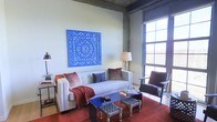 This single-floor space showcases a thoughtfully curated living area that blends industrial and bohemian design influences. The room is anchored by exposed concrete structural columns and high ceilings, lending a raw, loft-like character to the space. Large, grid-paned windows flood the interior with abundant natural light, offering views to the outdoors and creating an open, airy atmosphere.

The living area centers around a classic white tufted sofa styled with deep red and slate-blue accent pillows and a rich burgundy throw, complemented by a dark wood armchair with a woven seat nearby. A vibrant cobalt blue coffee table serves as a bold focal point on the rich red area rug, surrounded by woven ottomans that add texture and versatility.

The walls are kept in a neutral, muted tone, allowing the standout décor pieces to shine — most notably, a large, intricately carved cobalt blue wall panel that commands attention above the sofa. Warm ambient lighting from a classic table lamp softens the industrial edges of the space. Overall, the room balances bold color, artistic detail, and comfortable furnishings into a cohesive and inviting environment.