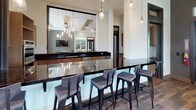 This single-story clubhouse-style space combines sophisticated design with functional amenities across several distinct areas. The lounge areas are defined by dramatically tall ceilings and floor-to-ceiling windows dressed in sheer white curtains, flooding the interiors with natural light and offering outdoor views. Cream-toned sofas, wingback chairs, and richly patterned area rugs create a warm, inviting atmosphere, while a wood-manteled fireplace with a wall-mounted television above adds a cozy focal point. An elegant chandelier with exposed bulbs anchors one seating area, enhancing the refined ambiance.

The kitchen is outfitted with light wood cabinetry extending to ceiling height, dark granite countertops, and stainless steel appliances including double wall ovens and a built-in microwave. Wood-look tile flooring runs throughout, adding warmth and cohesion. Adjacent to the kitchen, a sleek bar area features an expansive dark granite countertop, a stainless steel sink, and pendant lighting overhead. A pass-through window opens to the main gathering space, where a pool table and wall-mounted television are visible, making the space well-suited for entertaining and communal activities.