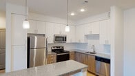 This thoughtfully designed one-bedroom, one-bathroom residence offers a clean, contemporary aesthetic across a single level. The open-concept kitchen serves as a central highlight, featuring stainless steel appliances including a refrigerator, range, microwave, and dishwasher. White upper cabinetry pairs with warm wood-toned lower cabinets, complemented by a subway tile backsplash and granite-look countertops. A peninsula island with pendant lighting above provides additional prep space and casual seating opportunities.

The living area flows naturally from the kitchen, finished with light hardwood-style flooring and recessed lighting that creates a bright, welcoming ambiance. A window brings in natural light, adding to the airy feel of the space.

The bedroom is carpeted for comfort and features a ceiling fan, natural light from dual windows with outdoor views, and direct access to both a walk-in closet and the full bathroom. The closet offers shelving and hanging space for ample storage.

The bathroom is finished with neutral large-format tile surrounding a full soaking tub and shower combination, maintaining the unit's clean, modern character throughout.