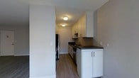 This single-floor space showcases a well-appointed galley-style kitchen as its central feature. The kitchen is designed with clean, modern aesthetics, highlighted by white flat-panel cabinetry that extends from floor to ceiling, maximizing storage capacity along both the upper and lower walls. Dark countertops provide a striking contrast against the white cabinetry, adding visual depth to the space.

Stainless steel appliances, including a refrigerator and dishwasher, contribute a sleek, contemporary feel. The undermount sink is positioned beneath a modern faucet along the countertop run, offering practical workspace. The kitchen's linear layout creates an efficient flow from one end to the other.

Wood-look flooring runs throughout, adding warmth to the otherwise neutral color palette of soft grey walls. Flush-mount ceiling light fixtures provide consistent illumination along the corridor-style layout. The textured ceiling adds a subtle architectural detail overhead. The overall ambiance is clean, functional, and modern, reflecting a thoughtfully designed interior suited for everyday living.