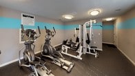 Located at 1272 Union Rd. in Gastonia, NC, this single-floor fitness center is designed to provide a well-equipped workout environment for its users. The room is finished with neutral gray walls accented by a bold teal horizontal stripe that runs continuously around the perimeter, creating a clean and energetic aesthetic. The flooring consists of durable, dark rubber gym matting, ideal for supporting heavy equipment and high-impact activity. Overhead, flush-mount ceiling lights provide consistent, even illumination throughout the space. A wall-mounted mirror spans a generous portion of one wall, enhancing the sense of space and allowing users to monitor their form during exercise. The facility is equipped with multiple elliptical machines, a treadmill, and a multi-station strength training machine, offering a comprehensive range of cardio and resistance workout options. A window fitted with white horizontal blinds allows for natural light and ventilation, while a fire safety pull station is mounted nearby for compliance and safety. Posted exercise room rules are clearly displayed on the wall. A pass-through window connects the fitness room to an adjacent, naturally lit space, suggesting proximity to a community room or leasing area. The overall layout is practical and functional, catering to everyday fitness needs.