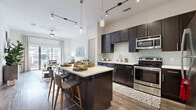 This single-story unit offers a thoughtfully designed open-concept layout that maximizes its compact footprint. The kitchen serves as a central focal point, showcasing dark espresso cabinetry with sleek hardware, light quartz countertops, a subway tile backsplash, and a full suite of stainless steel appliances. A multi-seat island with pendant lighting above connects the kitchen seamlessly to the dining and living areas, enhancing the sense of openness. Track lighting runs along the ceiling, supplementing the natural light that pours in from large sliding glass doors in the living room, which open to an outdoor balcony with exterior views. Wood-look flooring extends throughout the main living areas, adding warmth and continuity. The bedroom is carpeted and generously proportioned, featuring a large window that brings in natural light and outdoor views. It connects directly to a walk-in closet equipped with built-in shelving and hanging rods for organized storage. The bathroom features large-format tile flooring, full-height wall tile surrounding the soaking tub and shower combination, and crisp white panel doors throughout. Elevated ceilings contribute to the airy, modern ambiance of the overall space.