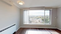 This single-floor residence is designed to maximize natural light and views, creating a bright and airy atmosphere throughout. The main living area is a standout feature, anchored by a large picture window that frames expansive views of the surrounding neighborhood's rooftops and lush tree canopy. Crown molding traces the perimeter of the room, adding a refined architectural detail, while rich hardwood flooring runs throughout, lending warmth and continuity to the space. A wall-mounted heating and cooling unit ensures year-round comfort. The compact kitchen is efficiently laid out with white cabinetry — including upper glass-front display cabinets — white countertops, and white appliances, including a range and dishwasher. A window above the sink allows natural light to brighten the workspace. The bathroom features a tiled tub-shower combination, a pedestal-style sink with brushed nickel fixtures, a mirrored medicine cabinet, and dark upper cabinetry for storage. A closet with built-in shelving provides additional organizational space. The unit's neutral gray walls and consistent hardwood flooring create a cohesive, clean aesthetic, while the elevated position of the living area offers particularly striking panoramic views of the tree-lined surroundings.