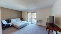 This single-floor unit offers a well-organized layout that maximizes functionality across its living spaces. The generously sized main bedroom is carpeted and bathed in natural light, featuring a textured ceiling and a warm neutral color palette with a contrasting accent wall. A sliding glass door opens directly onto a private balcony furnished with seating, offering pleasant outdoor views of surrounding greenery. A wall-mounted air conditioning unit ensures year-round comfort in this room.

The kitchen is efficiently designed with warm oak cabinetry providing ample storage, white appliances including a refrigerator, electric range, and microwave, and a countertop peninsula that doubles as a casual dining area with seating for two. The flooring transitions to easy-care tile in this space, and overhead lighting keeps the area bright and practical.

A secondary room, partially divided by a partial wall partition, serves as a flexible sleeping or study area. It features carpeted flooring, a work desk, and warm golden accent walls, creating a cozy and focused atmosphere. A floor-level heating vent ensures this space stays comfortable year-round.

The bathroom is clean and straightforward, appointed with a vanity featuring a stone-look countertop, a large mirror, and a shower/tub combination with a curtain enclosure. Together, these spaces form a compact yet complete and livable unit.