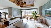 This single-floor space showcases a thoughtfully curated interior that merges bohemian warmth with contemporary sophistication. The open layout is anchored by rich, reclaimed hardwood flooring that flows throughout, punctuated by decorative tiled area rugs that define distinct zones within the space.

A standout feature is the coffered ceiling inset with reclaimed wood planking, complemented by striking woven pendant light fixtures that add an organic, artisanal quality. Botanical-patterned wallpaper in muted sage tones lines key accent walls, creating a lush, nature-inspired atmosphere that pairs beautifully with abundant live greenery throughout the space.

Floor-to-ceiling steel-framed windows and glass doors flood the interior with natural light while offering views to an adjacent outdoor terrace. Sheer white drapery panels soften the light and add an airy elegance to the space.

The layout accommodates multiple functional areas, including a relaxed lounge seating zone with a daybed and eclectic accent chairs, a central living area with a low concrete coffee table, and a dedicated dining or game area featuring a round stone-topped table surrounded by leather cane-back chairs. The overall ambiance is relaxed yet refined, with layered textures and carefully chosen furnishings contributing to a welcoming, design-forward environment.