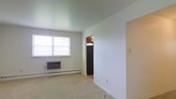 This single-floor residence offers a straightforward and functional layout designed for comfortable everyday living. The main living area is finished with wall-to-wall carpeting underfoot and smooth, light-painted walls that contribute to a clean and neutral aesthetic throughout. A standard window along one wall allows natural light to filter into the room, brightening the space during the day. A wall-mounted heater unit sits beneath the window, providing efficient climate control, while baseboard trim runs the perimeter of the room for a tidy finish. The ceiling is smooth and painted in a soft, muted tone that complements the overall palette of the interior. An open doorway leads from the living area directly into the kitchen, where a full-size refrigerator is visible, suggesting a practical and accessible kitchen layout. The transition between spaces is seamless, making the floor plan feel connected and easy to navigate. The overall ambiance of the space is calm and understated, presenting a blank canvas that is well-suited for personalization. The unit's single-floor design ensures convenience and ease of movement throughout.
