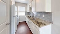 This single-floor space offers a clean, modern aesthetic defined by neutral gray walls, crisp white trim, and wood-look plank flooring that extends consistently throughout. The open living area is spacious and well-lit, featuring a ceiling fan with integrated lighting and a textured ceiling. Natural light filters in through multiple windows dressed with white horizontal blinds, creating a bright and airy atmosphere. The layout connects the living area to additional rooms via open doorways and a hallway, providing a functional flow throughout the unit.

The kitchen is a standout feature, showcasing granite countertops with bold veining, white shaker-style cabinetry with brushed nickel hardware, a double stainless steel undermount sink with a gooseneck faucet, and a light gray subway tile backsplash. A window above the sink brings in natural light.

The bedrooms are well-proportioned, each finished with the same wood-look flooring and neutral tones. One bedroom includes a window-mounted AC unit. The bathroom is efficiently designed with a white vanity, medicine cabinet mirror, brushed nickel fixtures, a toilet, and tile flooring, maintaining the clean, contemporary palette found throughout the space.