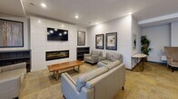 This well-appointed communal lounge space in California is designed to offer comfort and functionality across its generously sized single-floor layout. The open-plan interior is anchored by a striking feature wall housing a sleek, linear electric fireplace beneath a wall-mounted flat-screen television, framed by clean white paneling that lends a modern aesthetic. Multiple distinct seating arrangements are thoughtfully distributed throughout the space, including plush leather sofas, accent armchairs, and wooden coffee tables, creating inviting areas for both socializing and relaxation. Soft, neutral-toned carpeting runs throughout, complementing the warm beige walls and recessed lighting overhead. Large sliding glass doors and floor-to-ceiling windows along one wall admit an abundance of natural light while providing views to the exterior. A quieter nook tucked near the windows features a pair of leather armchairs flanked by tall potted plants, offering a more intimate setting. Decorative framed artwork is displayed throughout, adding visual interest and a polished finish to the overall ambiance. A stylish console table with a decorative mirror completes the space, reinforcing its role as a welcoming, communal gathering area.