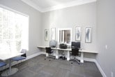 This single-floor space functions as a shared business center, thoughtfully designed to accommodate both focused work and casual collaboration. The room is finished with soft gray-toned walls, crisp white crown molding, and bold white baseboards that lend a polished, professional character throughout.

The workstation area lines one wall with a continuous light-wood desk surface, accommodating multiple desktop computers and a printer, each paired with ergonomic rolling chairs. Framed artwork featuring feather and nature-inspired prints adds a decorative touch above the workstations. A window fitted with white horizontal blinds allows controlled natural light into this area.

A dedicated meeting nook provides a small round white table with modern chairs, positioned beside a large window that fills the area with ample daylight. A wall-mounted shelf nearby offers additional workspace. A framed nature-inspired print decorates the adjacent wall.

The space is accessed through a glass-paneled door, and the noticeably tall ceilings contribute to an open, airy atmosphere. Patterned carpet tiles in a blend of tan, gray, and blue tones cover the floor throughout, adding texture while remaining functional. Overall, the space is well-organized and conducive to productivity.