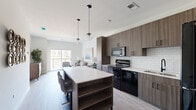 Located at 250 Plaza Circle in Mundelein, IL, this 2-bedroom, 1-bathroom residence offers a well-designed, single-floor layout with modern finishes throughout. The open-concept kitchen flows seamlessly into the living area, anchored by a large island with a white quartz countertop, built-in shelving, and bar seating. The kitchen features dark wood-grain cabinetry, a white subway tile backsplash, black appliances including a range, microwave, and refrigerator, and globe pendant lighting overhead. Light wood-style flooring runs throughout the main living areas, contributing to a clean, contemporary aesthetic. The living room is bright and airy, with sliding glass doors that open to a balcony offering outdoor views. The primary bedroom is generously sized, featuring plush carpet, a large window with outdoor views, and ample natural light. A carpeted hallway connects the bedrooms to the shared bathroom and closet. The bathroom is a standout feature, showcasing floor-to-ceiling marble-style tile, a glass-enclosed walk-in shower with a matte black frame, rainfall showerhead, and matte black fixtures throughout. A dedicated closet with built-in rod storage provides practical organization. The overall ambiance is modern, clean, and comfortable.
