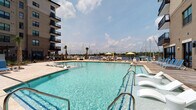 Located at 901 Nutt St in Wilmington, North Carolina, this outdoor amenity space offers a well-appointed, resort-style environment with direct views of a neighboring marina. At its centerpiece is a large, sparkling swimming pool flanked by lounge chairs and palm trees, creating a coastal atmosphere that takes full advantage of the waterfront setting. Sun umbrellas and dedicated sunbathing areas surround the pool deck, which is finished in clean, light-toned pavers.

Adjacent to the pool, a covered patio with floor-to-ceiling sliding glass doors connects seamlessly to interior spaces, blending indoor and outdoor living. The patio is outfitted with outdoor dining furniture and a mounted flat-screen television, making it ideal for gatherings. Stone and brick detailing on the building's exterior lend a refined architectural character throughout.

A dedicated grilling and outdoor kitchen area equipped with built-in gas grills and prep surfaces provides a functional space for outdoor cooking and entertaining. Nearby, a landscaped lawn area offers a relaxed setting with Adirondack chairs, a cornhole game, and a pergola-covered lounge with string lighting and ceiling fans. Stone-paved walkways connect each zone, tying the entire outdoor space together into a cohesive, community-oriented retreat.