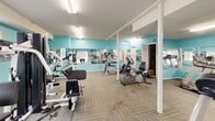 Located at 201 Queensbury Dr in Huntsville, Alabama, this dedicated fitness center offers a well-equipped exercise space designed to support a variety of workout routines. The room is finished with eye-catching teal accent walls that lend the space an energetic and motivating atmosphere, complemented by bright overhead fluorescent lighting that ensures excellent visibility throughout.

The floor is covered in multi-toned carpet tiles arranged in a geometric pattern, providing both comfort and durability underfoot. Large wall-mounted mirrors span multiple walls, visually expanding the space and allowing users to monitor their form during exercise.

The facility is outfitted with a comprehensive selection of equipment, including treadmills, a stationary upright bike, a recumbent bike, a multi-function cable strength machine, and additional cardio machines. This variety supports both cardiovascular and resistance training needs. A workout reference chart is posted on the wall, offering guidance for users of all fitness levels.

Natural light filters in through shuttered windows, and a set of double doors provides an additional access point, enhancing ventilation and connectivity. Structural columns are incorporated into the layout while maintaining an open, functional floor plan. The overall design is practical and purposeful, making this fitness center a convenient and well-rounded amenity.