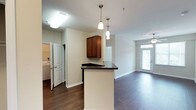 Located at 1001 Haynes Ln in Holly Springs, NC, this single-floor residence offers a well-planned open-concept layout designed for comfortable everyday living. Dark hardwood-style flooring flows throughout the main living areas, creating a warm and cohesive aesthetic.

The kitchen is a functional centerpiece, featuring rich medium-toned cabinetry, dark countertops, and black appliances including a range and over-the-range microwave. A peninsula with pendant lighting above provides a natural visual divide between the kitchen and the adjoining living area, while also offering additional counter space. Conveniently tucked just off the kitchen is a dedicated in-unit laundry area with a full-size washer and dryer.

The living area is generously proportioned and benefits from natural light streaming through a glass-paneled entry door, complemented by a ceiling fan overhead. The neutral wall palette throughout the home enhances the sense of openness.

The bedroom features plush carpeting, a ceiling fan, and a spacious closet with double doors and built-in rod storage. The bathrooms are finished with clean white tub-shower combinations, brushed nickel fixtures and hardware, and warm neutral walls. The overall ambiance of the space is clean, practical, and move-in ready, making it a solid residential option in Holly Springs.