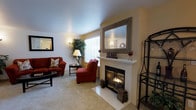 This single-floor residence offers a warm and inviting atmosphere highlighted by thoughtful design and comfortable living spaces. The living room serves as the centerpiece of the home, anchored by a classic white-mantled fireplace that provides both a functional heating element and a striking focal point. Above the mantle, a decorative framed mirror enhances the sense of space and reflects natural light throughout the room. The fireplace surround features clean tile work, adding a polished finishing touch.

The living area is finished with plush wall-to-wall carpeting that adds warmth underfoot, while smooth, light-colored walls and a bright white ceiling contribute to an open and airy feel. Large windows dressed with white blinds allow generous natural light to fill the room, further enhancing the bright and welcoming ambiance.

The space is well-proportioned, comfortably accommodating a full seating arrangement including a sofa and accent chair in rich red tones, complemented by dark wood side tables. Decorative touches such as potted plants, framed wall art, and accent lighting add character and personality throughout. Overall, this home presents a cozy, well-maintained living environment with classic design elements that provide both comfort and style.
