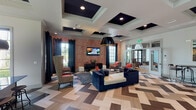 This sophisticated single-floor community lounge is a masterclass in contemporary interior design, blending bold aesthetics with comfortable, functional spaces. The entryway immediately impresses with a striking geometric-patterned floor — a mix of herringbone and diamond motifs in varied wood tones — that flows throughout the entire space, anchoring the eclectic décor. Soaring coffered ceilings with dark-painted insets add dramatic depth overhead, while oversized statement pendant lights in black and gold provide warm, focused illumination.

The main lounge area features a curated mix of seating, including plush velvet sofas, high-back accent chairs, and intimate conversation groupings arranged around patterned rugs. A boldly textured accent wall in warm rose tones serves as a focal point, complemented by a wall-mounted television and stylish side tables. Large floor-to-ceiling windows dressed with floor-length navy and charcoal curtains bathe the interiors in natural light while offering views of lush outdoor greenery, including palm trees.

A dedicated games area includes a full billiards table beneath dramatic pendant lighting, alongside a wet bar with mosaic tile backsplash and bar seating. A separate dining nook with a round stone-topped table and upholstered chairs provides a refined setting for smaller gatherings. An elegant arched entryway with a fanlight transom window adds a classic architectural touch, bridging the lounge's modern energy with timeless design.