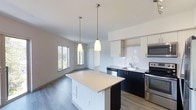 This two-bedroom, two-bathroom residence with a den presents a thoughtfully designed single-floor layout that maximizes both functionality and style. The open-concept living and kitchen area serves as the heart of the home, featuring sleek two-tone cabinetry with crisp white upper cabinets and bold dark lower cabinets, complemented by a classic subway tile backsplash. Stainless steel appliances, including a range and microwave, add a contemporary touch to the space.

A central kitchen island with a clean light-colored countertop provides additional prep space and serves as a natural gathering point, illuminated by two elegant pendant lights that hang overhead. The seamless flow from the kitchen into the living area creates a spacious, airy feel, enhanced by light wood-style flooring that runs throughout.

Large windows in the adjacent living space invite generous natural light and offer views of lush greenery outside, contributing to a bright and welcoming ambiance. The den provides flexible additional space suitable for a home office or study. Overall, the residence balances modern design with practical livability.