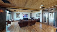 This single-floor space is thoughtfully designed to accommodate a variety of social and recreational activities, blending rustic and contemporary elements throughout. The main lounge area features soaring ceilings, crisp white walls, and abundant natural light filtering through large French doors that open to an outdoor area. A striking floor-to-ceiling stone fireplace with a solid wood mantle serves as a focal point, complemented by plush seating arranged for conversation and relaxation. Elegant chandelier lighting and neutral-toned furnishings, including a cream sofa and wingback chairs, create a welcoming, refined atmosphere.

An adjoining media room offers a distinctly different character, showcasing a warm tongue-and-groove wood-paneled ceiling, rich hardwood flooring, and recessed lighting. Vintage boxing and entertainment posters adorn the white walls, adding a bold, themed personality to the space. Deep brown leather sofas and armchairs are arranged around a large flat-screen television, making it ideal for group viewing. A billiards table is partially visible, further enhancing the recreational appeal.

Together, the areas balance comfort and character, offering a well-rounded environment suitable for both casual gatherings and leisure activities.