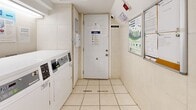 Located at 223 Woodbine Ave in Toronto's East End, this shared laundry facility is a functional and well-organized amenity space. The room features a clean, utilitarian design with white tile flooring and white-painted walls throughout, creating a bright and tidy environment. A row of modern front-loading washing machines lines one side of the space, accompanied by matching dryers, offering ample capacity for residents. A deep utility sink is positioned along the wall, providing a convenient station for pre-treating garments or handwashing items. Fluorescent strip lighting runs along the ceiling, ensuring the space is well-lit from end to end. Informational signage, including resident notices and usage instructions, is posted throughout, keeping users informed of guidelines and procedures. A white-paneled door provides access to the building, while a tiled staircase at the rear of the room connects to upper floors, flanked by utility infrastructure including wiring and storage. The overall layout is efficiently arranged to maximize usability within a compact footprint, making this laundry room a practical and accessible shared resource for building residents.