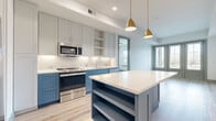 This single-floor unit showcases a thoughtfully designed interior with a cohesive modern aesthetic. The kitchen serves as a standout feature, distinguished by its two-tone cabinetry — crisp white upper cabinets contrast with slate blue lower cabinets, all topped with bright white countertops. A central island with open shelving provides additional storage and prep space, while gold pendant lights overhead add a warm, contemporary accent. Stainless steel appliances, including a French door refrigerator, range, microwave, and dishwasher, complete the well-appointed kitchen. Light wood-look flooring runs consistently throughout the unit, complemented by sage green baseboards and door trim that add a subtle, distinctive detail to each room. The living and bedroom areas are bright and open, benefiting from natural light through windows dressed with horizontal blinds, and a ceiling fan enhances comfort in the bedroom. The bathroom offers a striking design contrast, featuring warm terracotta hexagonal floor tiles paired with a blue-grey vanity, white subway tile shower surround, a soaking tub, and a large vanity mirror with modern sconce lighting. The overall ambiance is clean, fresh, and move-in ready.