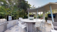 This sophisticated outdoor entertaining space showcases a striking curved architectural design, with smooth concrete columns and walls that frame the area with a sense of refined structure. At the heart of the space sits a generously sized round granite-topped bar counter, surrounded by white slatted bar-height chairs, making it an ideal gathering spot for guests. The polished stone tile flooring adds a clean, upscale aesthetic underfoot.

A built-in stainless steel grill is conveniently positioned along the curved wall, making outdoor cooking effortless, while a wall-mounted outdoor television enhances the entertainment experience. The pavilion-style design allows for open-air enjoyment while providing a sense of enclosure and intimacy.

The space is enveloped by dense, mature tropical landscaping, including tall palms and abundant greenery that create a private, resort-like atmosphere. The lush foliage serves as a natural backdrop, adding vibrant color and texture while offering shade and seclusion. The open sky above fills the area with natural light, contributing to the bright and airy ambiance. Overall, this outdoor space offers a seamless blend of functional amenities and tropical elegance, ideal for both casual relaxation and entertaining.
