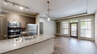 Located at 1460 Distribution Dr in Suwanee, GA, this single-floor residence offers a well-designed, open-concept layout that balances functionality with modern style. The kitchen serves as the heart of the home, showcasing a granite-topped island with a double stainless steel sink, rich dark wood cabinetry, stainless steel appliances including a refrigerator, range, and microwave, and elegant pendant lighting above the island complemented by track lighting. The kitchen flows seamlessly into the living area, which features warm hardwood-style flooring, crown molding, a tray ceiling, built-in shelving, and large windows and a glass door that invite generous natural light and outdoor views. An arched doorway adds a classic architectural touch connecting spaces throughout the unit. The bedroom is a comfortable, well-lit retreat with hardwood-style flooring, a ceiling fan, and a broad window overlooking greenery. A spacious walk-in closet with wire shelving and dedicated laundry hookups provides excellent storage and convenience. The bathroom is finished with tile flooring, a granite vanity countertop, and a full tub-shower combination with white tile surround. Throughout the residence, neutral tones and consistent finishes create a cohesive and inviting atmosphere.