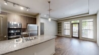 Located at 1460 Distribution Dr in Suwanee, GA, this single-floor residence offers a well-designed, open-concept layout that balances functionality with modern style. The kitchen serves as the heart of the home, showcasing a granite-topped island with a double stainless steel sink, rich dark wood cabinetry, stainless steel appliances including a refrigerator, range, and microwave, and elegant pendant lighting above the island complemented by track lighting. The kitchen flows seamlessly into the living area, which features warm hardwood-style flooring, crown molding, a tray ceiling, built-in shelving, and large windows and a glass door that invite generous natural light and outdoor views. An arched doorway adds a classic architectural touch connecting spaces throughout the unit. The bedroom is a comfortable, well-lit retreat with hardwood-style flooring, a ceiling fan, and a broad window overlooking greenery. A spacious walk-in closet with wire shelving and dedicated laundry hookups provides excellent storage and convenience. The bathroom is finished with tile flooring, a granite vanity countertop, and a full tub-shower combination with white tile surround. Throughout the residence, neutral tones and consistent finishes create a cohesive and inviting atmosphere.