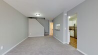 This two-floor space offers a clean, functional layout with a neutral color palette that creates a calm and welcoming atmosphere throughout. The main level features an open living area finished with soft, light gray carpeting, providing a comfortable and versatile space. Pale gray walls and white trim contribute to a bright, cohesive aesthetic that carries through the entire floor.

A staircase leads to the upper level, with the understair area neatly integrated into the living space. Adjacent to the living area, the kitchen is accessible through an open pass-through and a wide doorway, offering seamless connectivity between the two spaces. The kitchen is equipped with stainless steel appliances, including a full-size refrigerator, and features white cabinetry that provides ample storage. The kitchen floor transitions to warm hardwood-style flooring, adding a layer of contrast and texture to the space.

Flush-mount ceiling lighting illuminates both the living area and the kitchen, ensuring consistent brightness throughout. The overall design is practical and understated, with a focus on clean lines and efficient use of space. The two-floor layout offers room for a variety of living arrangements, making this a versatile and well-appointed space.