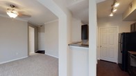 This single-floor residence offers a well-organized layout that balances functionality and comfort. The interior is defined by neutral, warm-toned walls that create a bright and welcoming atmosphere throughout. A notable architectural feature is the open pass-through kitchen bar, framed by structural columns and topped with a dark granite-style countertop, providing a natural division between the kitchen and the adjacent living areas while maintaining an open feel. The kitchen is equipped with dark espresso-toned cabinetry, lending a modern contrast to the light-colored walls. Flooring transitions thoughtfully between dark hardwood in the kitchen and entryway areas and plush, light-toned carpet in the living and bedroom spaces, adding warmth and texture underfoot. Crown molding runs along the ceiling, providing a refined architectural detail that elevates the overall design. Doorways throughout are finished with classic panel-style doors and white trim, contributing to a clean, cohesive aesthetic. The layout allows for smooth circulation between rooms, with corridors leading to what appear to be a bedroom and additional living spaces. Overall, this unit presents a practical and comfortable living environment with thoughtful design details.