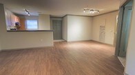 This single-floor space offers a clean, open layout designed for both comfort and functionality. The interior is finished with warm hardwood-style flooring that runs throughout, complementing the neutral, light-toned walls and creating a welcoming ambiance. A striking track lighting fixture on the ceiling provides focused illumination, enhancing the brightness of the space alongside natural light that filters in through a window near the kitchen area. A raised counter or bar-height partition subtly defines the transition between the kitchen area and the main living space, adding a sense of structure without compromising the open feel. The entry is anchored by a classic paneled door in a contrasting gray-green tone, adding a touch of character to the otherwise understated interior. Crown molding detail along the upper walls adds a refined architectural touch throughout. The spacious main area is highly versatile and well-suited for a variety of furniture arrangements and uses. Overall, the space presents a tidy, move-in-ready environment with a neutral palette that provides an excellent canvas for personalization.