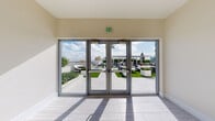 This rooftop-level vestibule serves as a transitional space connecting the building's interior elevator lobby to an expansive outdoor terrace. The interior is finished in a clean, contemporary style, with light wood-toned tile flooring and smooth, neutral-toned walls accented by crisp white baseboards. A stainless steel elevator with polished doors provides vertical access, and safety signage is mounted neatly on the adjacent wall.

Floor-to-ceiling glass doors with aluminum frames span the exterior-facing wall, flooding the space with abundant natural light and providing clear sightlines to the rooftop terrace beyond. The outdoor area visible through the glass features manicured artificial turf, large white planters with greenery, and multiple seating arrangements including lounge chairs and a pergola-covered bar area. Tiled pathways connect the various outdoor zones, and the surrounding urban skyline, including nearby buildings and palm trees, frames the backdrop under open skies.

The vestibule's understated, minimal design prioritizes the visual connection to the outdoor amenity space, creating a seamless indoor-to-outdoor transition that is both functional and inviting.