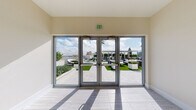 This rooftop-level vestibule serves as a transitional space connecting the building's interior elevator lobby to an expansive outdoor terrace. The interior is finished in a clean, contemporary style, with light wood-toned tile flooring and smooth, neutral-toned walls accented by crisp white baseboards. A stainless steel elevator with polished doors provides vertical access, and safety signage is mounted neatly on the adjacent wall.

Floor-to-ceiling glass doors with aluminum frames span the exterior-facing wall, flooding the space with abundant natural light and providing clear sightlines to the rooftop terrace beyond. The outdoor area visible through the glass features manicured artificial turf, large white planters with greenery, and multiple seating arrangements including lounge chairs and a pergola-covered bar area. Tiled pathways connect the various outdoor zones, and the surrounding urban skyline, including nearby buildings and palm trees, frames the backdrop under open skies.

The vestibule's understated, minimal design prioritizes the visual connection to the outdoor amenity space, creating a seamless indoor-to-outdoor transition that is both functional and inviting.
