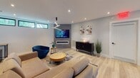 This thoughtfully designed single-floor space blends modern aesthetics with functional versatility. Light hardwood flooring runs throughout, tying together each distinct area while creating a cohesive, airy atmosphere.

The main lounge area serves as the heart of the space, featuring recessed lighting, a bold geometric chandelier, and plush blue velvet chairs arranged around a large decorative wall clock — ideal for welcoming guests or encouraging relaxed conversation. A built-in beverage station with dark cabinetry, open shelving, and a coffee setup offers a convenient refreshment hub.

A cozy media and seating area showcases a wall-mounted flat-screen TV above a sleek linear fireplace framed in stone tile. A sculptural pendant light fixture anchors the room, which is complemented by a sectional sofa, round wood coffee table, and curated wall art. Horizontal transom windows allow natural light to filter in.

An industrial-style shelving unit with wood and black metal frames separates the lounge from a dedicated fitness area, which is enclosed by glass panels and equipped with professional-grade exercise machines. The open-plan layout visible in the overhead view reveals a well-organized flow between the lounge, a treatment or service room, and common areas — making this space well-suited for a wellness center, boutique studio, or professional office environment.