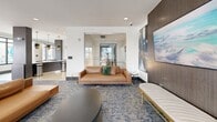 Located at 1213 Avalon Blvd in Alpharetta, Georgia, this well-appointed amenity lounge offers a sophisticated and versatile gathering space spread across a single floor. The interior showcases a contemporary design aesthetic, blending warm tones with bold accents through a carefully curated mix of materials and furnishings.

At the heart of the space is a striking curved bar island topped with a white quartz countertop and surrounded by sleek charcoal and gold-accented bar seating. Dramatic black and gold pendant lights hang overhead, complementing a geometric patterned tile backsplash. Adjacent to the bar, a full kitchenette provides added functionality with appliances and ample counter space.

The lounge area features plush leather sofas, emerald green velvet armchairs, and chaise seating arranged atop a richly patterned blue carpet. A billiards table anchors the central gathering zone, while multiple seating vignettes accommodate both social and relaxed use. Curated artwork lines the walls throughout, adding an art-gallery quality to the space.

Large, multi-pane windows flood the interior with natural light and offer views to an outdoor patio area. A glass-railed mezzanine overlook adds architectural depth, while textured accent wallcoverings, wood-paneled columns, and wall-mounted televisions complete the polished, modern atmosphere.
