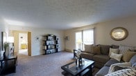 This single-floor residence offers a welcoming living room as its central gathering space. The room features wall-to-wall carpeting in a neutral tone, contributing to a warm and comfortable atmosphere. A textured ceiling adds subtle visual interest overhead, while light-colored walls keep the space feeling bright and open.

Natural light filters into the room through a large window dressed with sheer white curtains, offering a view to the exterior and enhancing the airy feel of the space. A wall-mounted air conditioning unit is conveniently positioned beneath the window, ensuring year-round comfort. A baseboard heater runs along the wall, providing an additional climate control option.

The living area is furnished with a plush, earth-toned sofa accented with decorative pillows, paired with a dark-framed glass-top coffee table. A striking open shelving unit mounted along one wall serves as a functional display area for decorative items, plants, and personal accents, adding character to the room. A door visible along the adjacent wall provides access to other areas of the home, suggesting a practical and well-connected floor plan throughout.