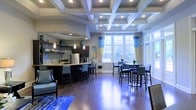 This single-floor space showcases an open-concept layout that seamlessly connects a kitchen, bar seating area, and dining space. The most striking architectural feature is the elaborate coffered ceiling, detailed with white-painted beams arranged in a geometric grid pattern and recessed lighting, lending a sense of refined structure overhead. Rich, dark hardwood flooring runs throughout, providing warm contrast to the bright, airy atmosphere.

The kitchen area features dark cabinetry with a tiled backsplash, stainless steel appliances, and a granite-topped island with bar-height seating. Pendant lights hang above the island, adding focused illumination. Adjacent to the kitchen, a comfortable lounge seating area with upholstered furniture and a blue area rug offers a relaxed gathering space.

A large bay window with blue and yellow curtains dominates one wall, flooding the dining area with natural light and offering views of surrounding greenery. The dining area accommodates a full table with multiple chairs. The overall color palette — navy blue accents, crisp white ceilings, and warm wood tones — creates an inviting and well-coordinated ambiance throughout the space.