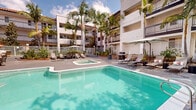 Located at 1657 Huntington Dr in Duarte, California, this outdoor recreational area showcases a well-appointed, resort-style courtyard designed for relaxation and leisure. The centerpiece is a generously sized swimming pool with clear turquoise water, accessible via stainless steel entry rails and surrounded by a smooth concrete deck ideal for sunbathing. Adjacent to the main pool sits a separate spa, offering a dedicated space for relaxation. Tall, mature palm trees are planted throughout the courtyard, providing both shade and a tropical ambiance that complements the sunny Southern California setting. An outdoor dining and lounge area is positioned nearby, furnished with tables, chairs, and vibrant patio umbrellas, making it well-suited for casual gatherings. Chaise lounge chairs are arranged around the pool deck for sunbathing. A gas grill station adds outdoor cooking convenience. The space is enclosed by a white iron fence, ensuring privacy and security. The surrounding multi-story building features a Spanish Colonial-inspired architectural style, with stucco exteriors, clay tile rooflines, and private balconies overlooking the courtyard. Mature shade trees further enhance the lush, green atmosphere of this inviting outdoor retreat.
