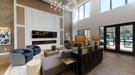 This thoughtfully designed single-floor community space combines contemporary elegance with functional comfort across several distinct yet interconnected zones. The centerpiece is a grand lounge area distinguished by soaring double-height ceilings, a coffered ceiling detail, and a striking linear fireplace set into a floor-to-ceiling feature wall of paneled millwork and warm wood accents. A large flat-screen display is mounted above the fireplace, complemented by a dramatic multi-globe chandelier that casts a warm, ambient glow over the generously scaled seating arrangement. Expansive floor-to-ceiling windows and clerestory glazing invite abundant natural light and frame views of the surrounding outdoor areas, including what appears to be a resort-style pool courtyard. Wide-plank hardwood flooring flows seamlessly throughout, unifying the open-concept layout. The kitchen area is a standout feature, offering an oversized marble-topped island with seating for multiple guests, globe pendant lighting with brushed brass fixtures, white cabinetry, quartz countertops, a textured tile backsplash, and premium stainless steel appliances. Adjacent to the kitchen, a semi-private dining nook with a marble-top round table and black steel-framed glass partition walls provides an intimate setting for smaller gatherings. Curated artwork, lush greenery, and carefully selected furnishings complete the polished, upscale ambiance throughout.