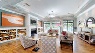 Located at 3868 Central Pike in Nashville, TN, this well-appointed community clubhouse offers a welcoming and versatile gathering space designed with both comfort and style in mind. The single-floor layout flows openly, anchored by rich hardwood flooring that runs throughout and tray ceilings with crown molding that lend an elevated sense of architecture.

The centerpiece of the main living area is a classic white-mantled fireplace flanked by built-in shelving stocked with decorative firewood, adding warmth and visual character. Nashville-inspired guitar artwork in bold orange tones punctuates the walls, nodding to the city's musical heritage. Multiple seating arrangements—featuring neutral upholstered sofas, patterned accent chairs, and coordinating ottomans with red accent pillows—create an inviting, lounge-like atmosphere.

Floor-to-ceiling sliding glass doors and large windows line one wall, flooding the space with natural light and offering direct views of an outdoor pool area complete with lounge seating and red umbrellas. Track lighting and statement pendant fixtures provide layered illumination throughout.

An adjacent dining nook with counter seating, warm wood cabinetry, and a wall-mounted TV completes the space, making it ideal for casual gatherings and community events alike.