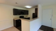 This single-floor space showcases a contemporary kitchen as its central feature. The kitchen is outfitted with sleek, dark espresso-toned cabinetry that extends to the upper walls, providing ample storage. The upper cabinets are complemented by matching lower cabinetry fitted with brushed silver hardware, creating a cohesive and modern aesthetic. Black appliances, including a range, microwave, and refrigerator, are seamlessly integrated into the design.

A peninsula countertop with a light-toned surface serves as a functional divider between the kitchen and the adjacent living space. This peninsula provides additional workspace and could serve as a casual dining or entertaining area. The countertops contrast effectively with the dark cabinetry, adding visual balance to the space.

The flooring transitions from a lighter carpet in the kitchen area to dark hardwood or laminate in the adjoining room, subtly defining each zone. Overhead, a flush-mount ceiling light provides direct illumination throughout the kitchen. The walls are finished in a neutral off-white tone, contributing to a clean and open atmosphere throughout the space.