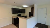 This single-floor space showcases a contemporary kitchen as its central feature. The kitchen is outfitted with sleek, dark espresso-toned cabinetry that extends to the upper walls, providing ample storage. The upper cabinets are complemented by matching lower cabinetry fitted with brushed silver hardware, creating a cohesive and modern aesthetic. Black appliances, including a range, microwave, and refrigerator, are seamlessly integrated into the design.

A peninsula countertop with a light-toned surface serves as a functional divider between the kitchen and the adjacent living space. This peninsula provides additional workspace and could serve as a casual dining or entertaining area. The countertops contrast effectively with the dark cabinetry, adding visual balance to the space.

The flooring transitions from a lighter carpet in the kitchen area to dark hardwood or laminate in the adjoining room, subtly defining each zone. Overhead, a flush-mount ceiling light provides direct illumination throughout the kitchen. The walls are finished in a neutral off-white tone, contributing to a clean and open atmosphere throughout the space.