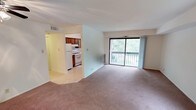 This single-floor unit is organized around a spacious, carpeted living and dining area that serves as the central hub of the layout. A large sliding glass door dominates one wall, providing direct access to a private balcony and framing views of surrounding greenery. An additional window further enhances natural light throughout the main living space, creating a bright and open atmosphere.

The kitchen is accessible directly from the living area and features rich, warm-toned wooden cabinetry that extends to the upper walls for ample storage. A gas range with overhead ventilation, a white refrigerator, and a stainless steel sink with a window above complete the functional cooking space. Light-toned countertops contrast with the darker cabinetry, providing a practical work surface.

The bedrooms are carpeted and well-proportioned, each with windows that bring in natural light and views of the outdoors. One bedroom includes a dedicated walk-in closet, offering generous storage, while another features a ceiling fan and additional closet space.

The bathroom is clean and efficiently designed, with a full tub and shower combo finished in white tile, a large vanity mirror with overhead lighting, and a single-sink vanity with storage beneath. The unit's neutral color palette and consistent carpeting throughout the living spaces contribute to a cohesive, understated aesthetic.